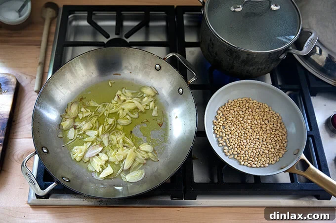 A kitchen stovetop scene showing three key elements: a large skillet gently sweating shallots and garlic in olive oil, a smaller skillet toasting pine nuts over low heat, and a pot of water coming to a boil for the pasta, all contributing to the preparation of a simple cherry tomato pasta.
