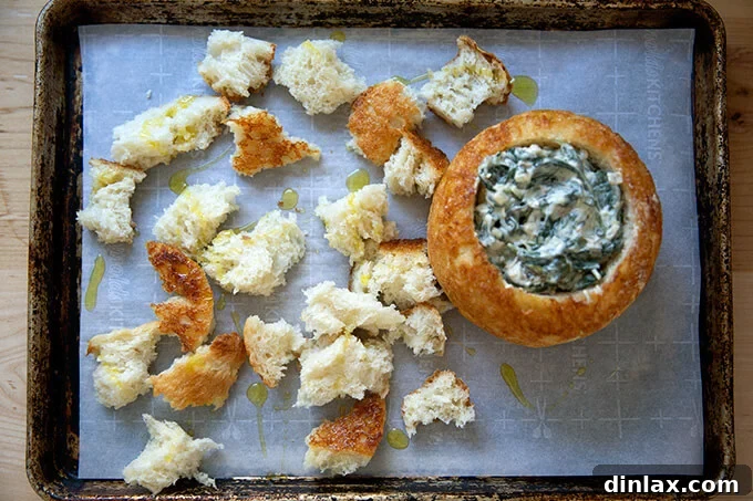 A sheet pan with a peasant bread bread bowl filled with spinach-artichoke dip, surrounded by toasted bread pieces.