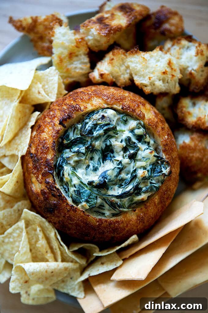 Spinach-artichoke dip served in a rustic bread bowl, garnished with fresh herbs, surrounded by crackers and vegetables.