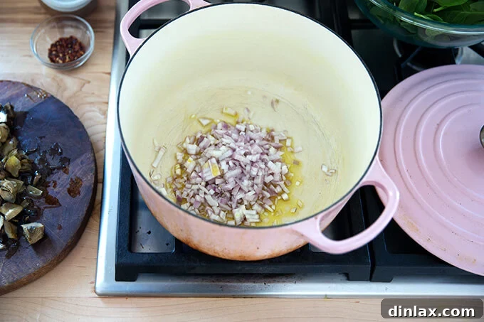 A large pot on a stovetop, containing olive oil, diced shallots, and a pinch of salt, ready to be cooked.