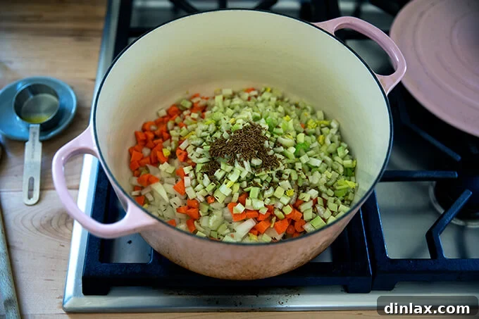 A large pot on the stovetop filled with diced onions, celery, carrots, and whole cumin seeds, gently sautéing in olive oil to create an aromatic base for the soup.