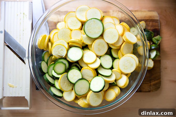 Sliced zucchini and summer squash in a large bowl next to a mandoline, ready for preparation.