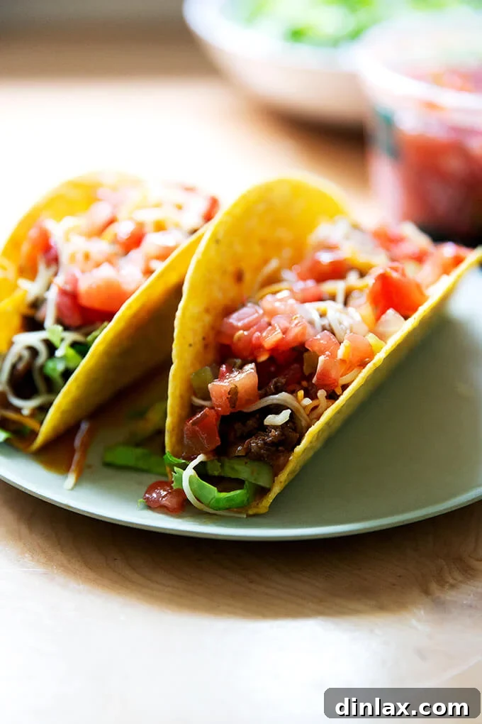 Two beautifully assembled hard shell tacos, generously filled with seasoned taco meat, fresh lettuce, shredded cheese, and a dollop of vibrant salsa, presented on a clean white plate.