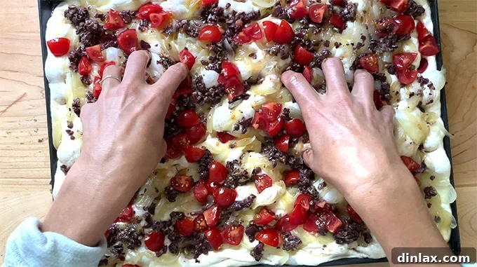 Hands gently dimpling the unbaked Pissaladière dough with its toppings already applied, prior to baking.