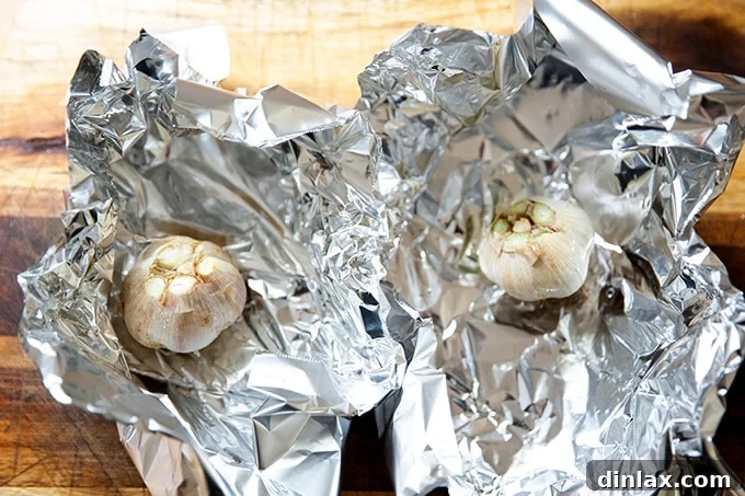 Two trimmed garlic heads, each on a small sheet of aluminum foil, drizzled with olive oil and water, ready for enclosure.