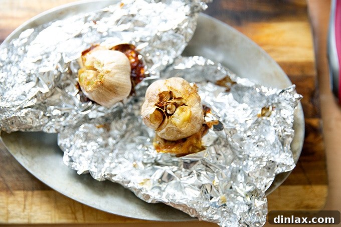 Roasted garlic heads, removed from the oven, with the foil pouches opened to reveal soft, golden, caramelized cloves.