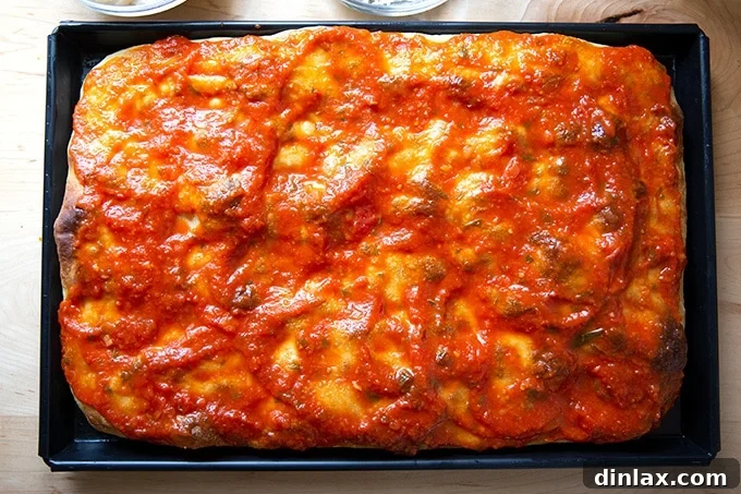 A parbaked Sicilian pizza crust generously coated with a vibrant red tomato sauce, ready for cheese.
