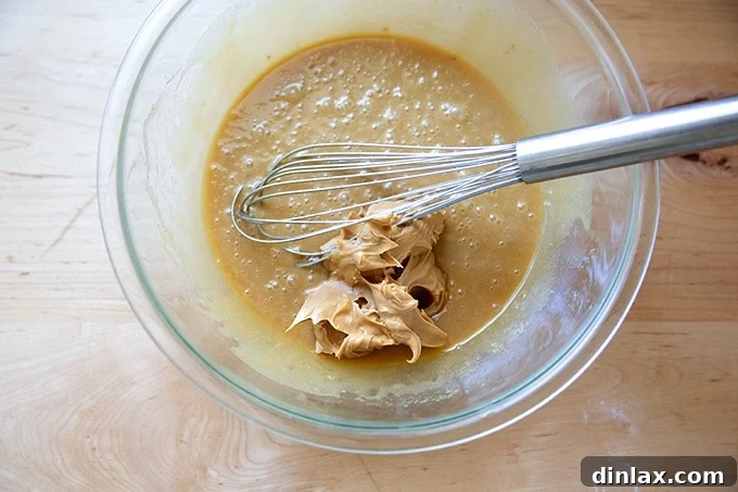 A large mixing bowl containing the developing peanut butter cookie dough, with peanut butter being vigorously mixed in with a whisk to create a smooth, unified batter.