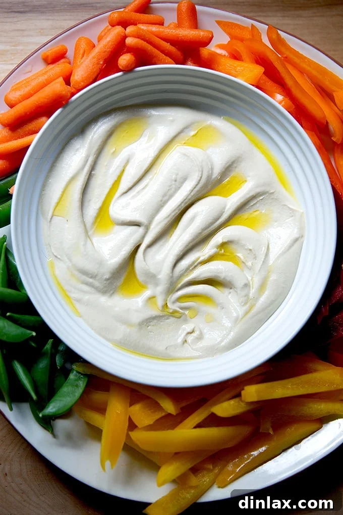A platter of creamy homemade hummus served with an assortment of fresh, colorful vegetables like bell peppers, carrots, and snap peas, garnished with a drizzle of olive oil.