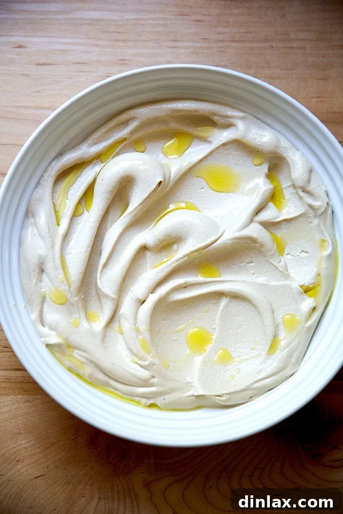 A bowl of exceptionally creamy hummus, made with a Vitamix, beautifully drizzled with olive oil and ready for garnishing, presented on a plate.