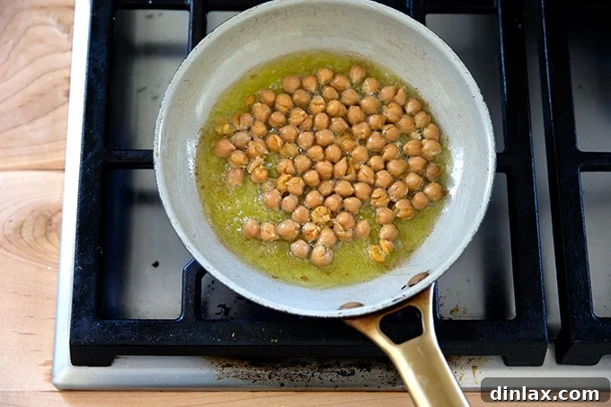 A small skillet shows chickpeas frying in olive oil, beginning to crisp and brown, on a stovetop.