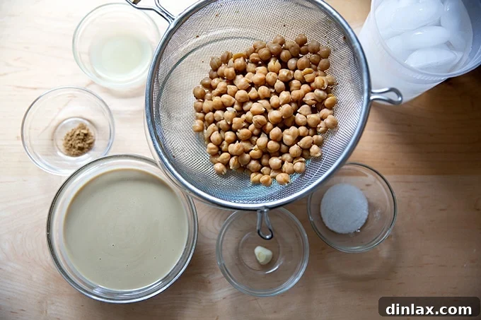 An overhead view of the simple ingredients for homemade hummus laid out on a clean countertop: tahini, cooked chickpeas, fresh lemons, and a bowl of salt.