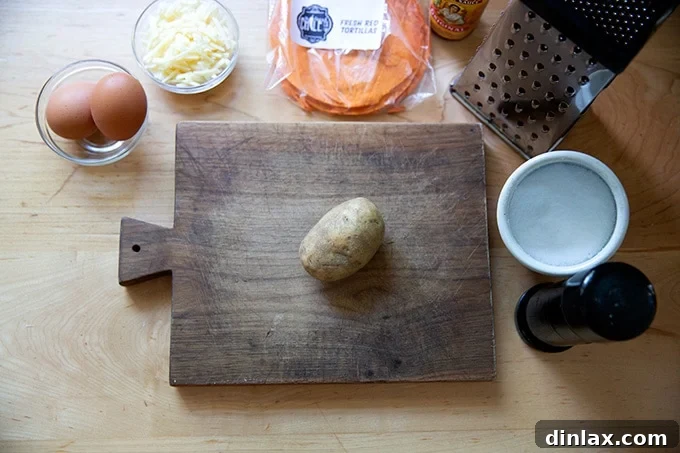 Essential ingredients for crispy potato tacos laid out: a small potato, eggs, a block of cheese, tortillas, salt, pepper, and a box grater on a wooden cutting board.