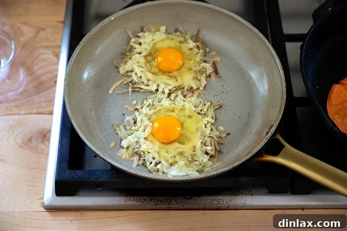 A close-up of a skillet showing two portions of crispy potato and cheese, each with a freshly cracked, uncooked egg nestled in its center.