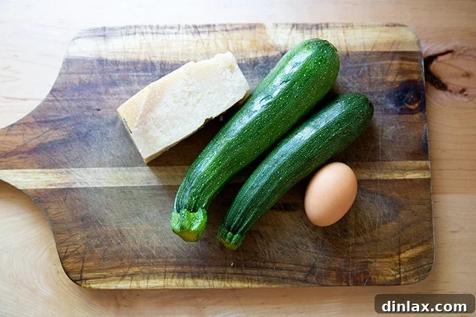 A block of parmesan, two zucchini, and an egg on a cutting board.