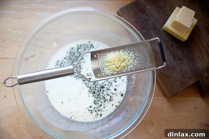 A microplane grater rests on the edge of a large mixing bowl, with finely minced garlic visibly gathered on its surface.