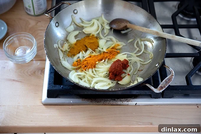 Close-up of a skillet on the stovetop showing sautéed onions mixed with bright yellow turmeric, curry powder, and a dollop of red Thai curry paste, expertly blended.