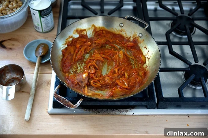A skillet on the stovetop, now containing sautéed onions, spices, and Thai red curry paste, with crushed tomatoes just added and being stirred in, creating a rich base.