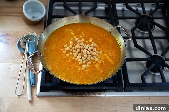 The skillet on the stovetop, now holding two cans of drained chickpeas stirred into the simmering curried sauce, ready for further cooking and flavor infusion.
