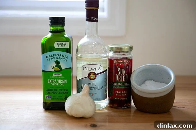 Ingredients for sun-dried tomato dressing laid out on a kitchen countertop: olive oil, white balsamic vinegar, sun-dried tomatoes, salt, and garlic clove.