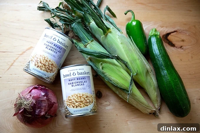 An assortment of fresh salad ingredients on a countertop: a whole red onion, two cans of white beans, two ears of corn, one green jalapeño, and one zucchini.