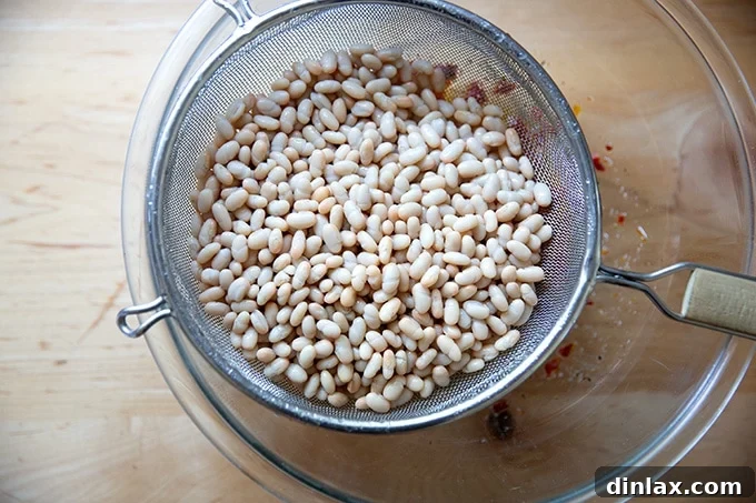White beans in a metal strainer, draining over a large glass bowl on a countertop.