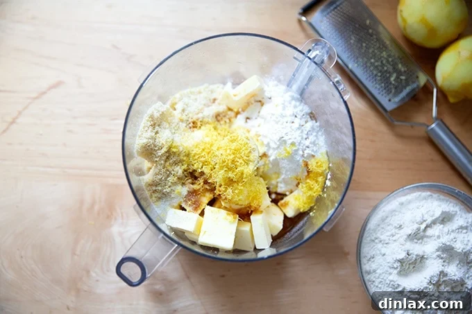 A food processor bowl filled with cold butter cubes, confectioners' sugar, lemon zest, vanilla, salt, and almond flour, before blending.