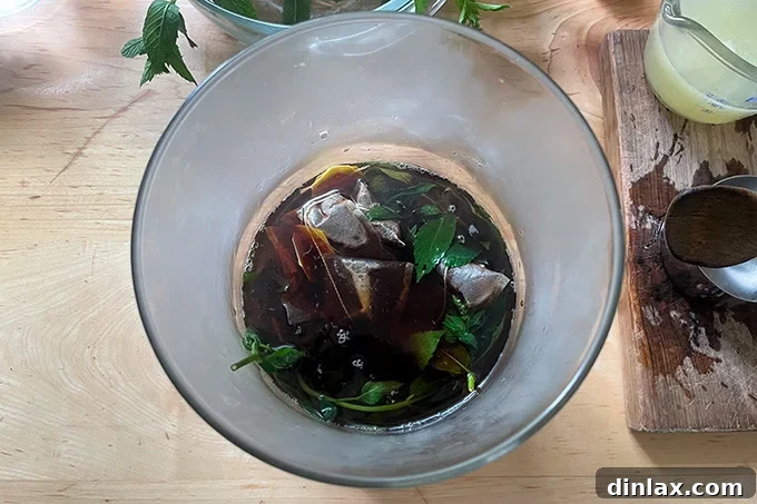 A glass vessel holding tea bags and fresh mint steeping in hot, clear water, with steam gently rising.