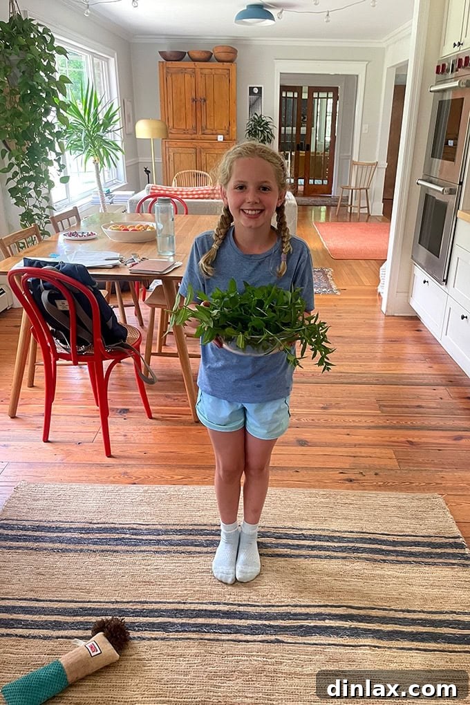 A young girl, Tig, proudly holding a bowl brimming with freshly picked green mint leaves, ready for iced tea preparation.