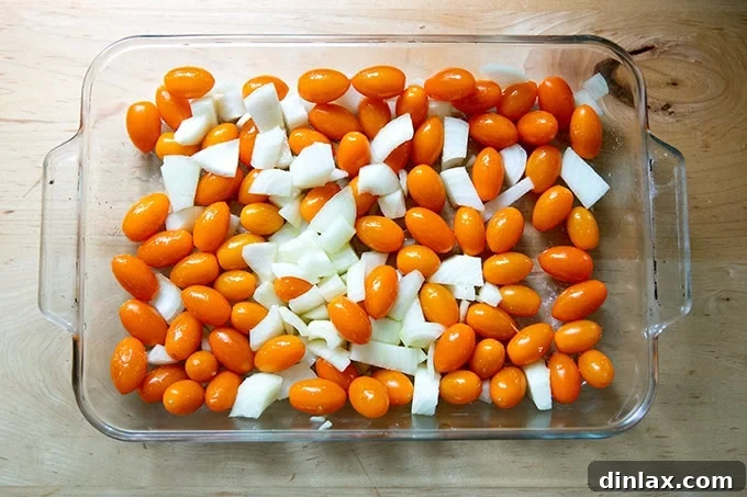 A close-up view of a 9x13-inch baking pan, showcasing vibrant yellow cherry tomatoes and chopped onions, glistening from being tossed with olive oil and salt, ready for the oven.