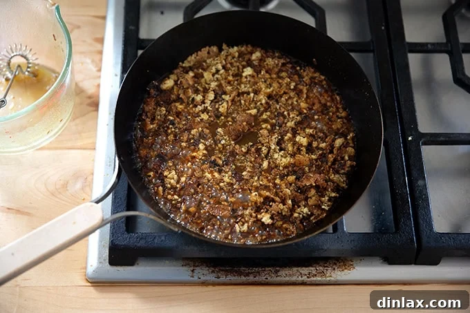 A skillet featuring crisped tofu crumbles being actively coated and saturated by the added miso-lime sauce, showing the rapid absorption.