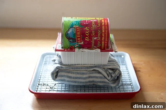 A cloth-wrapped block of tofu being pressed beneath a heavy can of tomatoes, resting on a cooling rack to drain excess liquid.