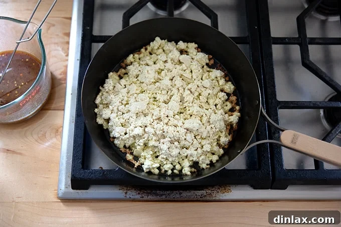 A hot skillet containing shimmering oil and a layer of fresh tofu crumbles, just beginning to cook.