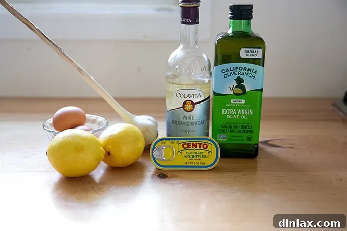 Perfect Caesar Dressing Your Way 4 Ingredients for Caesar dressing laid out on a countertop: fresh lemons, garlic cloves, anchovy tin, eggs, white balsamic vinegar, and a bottle of extra-virgin olive oil.