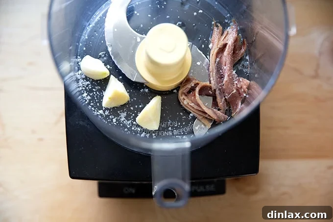 Perfect Caesar Dressing Your Way 5 Garlic cloves, flaky sea salt, and anchovies placed inside a food processor bowl, ready for blending.