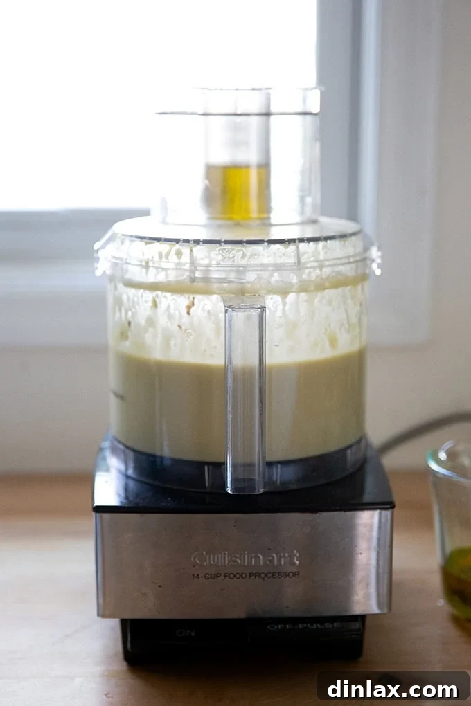 Perfect Caesar Dressing Your Way 8 A food processor with olive oil being slowly streamed into the mixture through its food chute, indicating the emulsification process.