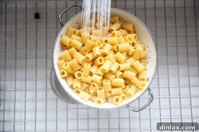 Effortless Baked Pasta: Make It, Share It, Freeze It 7 Pasta being rinsed with cold water in a colander within a kitchen sink.