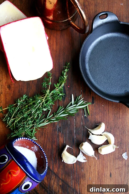 Ingredients for baked fontina laid out on a wooden board: Fontina cheese, fresh rosemary, fresh thyme, garlic cloves, olive oil, salt, and pepper. All components are fresh and ready for preparation.