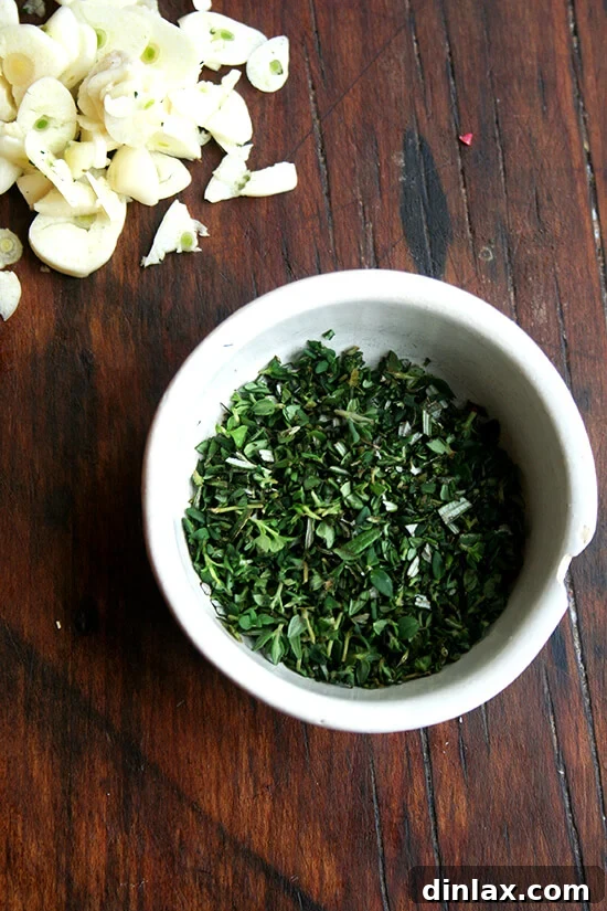 Freshly minced rosemary and thyme with thinly sliced garlic cloves on a cutting board, prepared for infusion into the baked fontina.