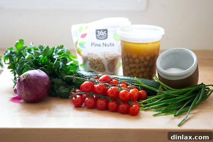 A display of fresh ingredients for the herby chickpea salad: cooked chickpeas, cherry tomatoes, cucumbers, red onion, bunches of parsley and chives, and a small bowl of pine nuts.