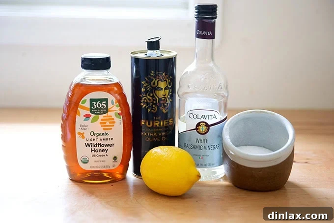 The ingredients for the lemon vinaigrette: a bottle of honey, olive oil, white balsamic vinegar, a lemon, and a container of salt, arranged on a kitchen countertop.