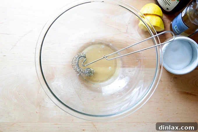 A large glass bowl containing honey, white balsamic vinegar, fresh lemon juice, and salt, being whisked together to form the base of the dressing.