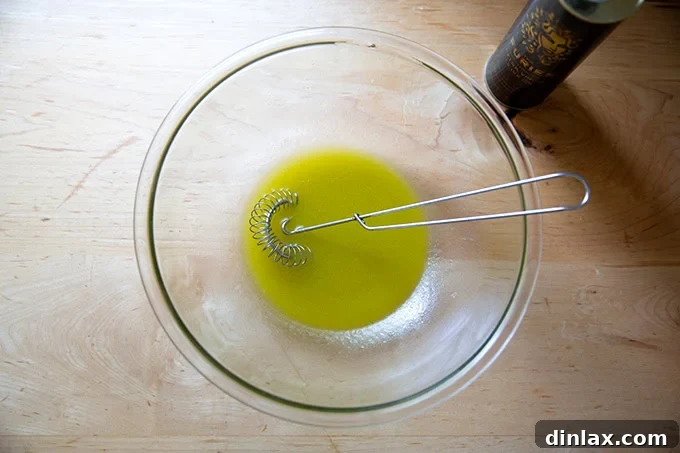 Extra-virgin olive oil being slowly streamed into a large bowl containing the whisked honey, white balsamic vinegar, lemon juice, and salt, creating an emulsified dressing.