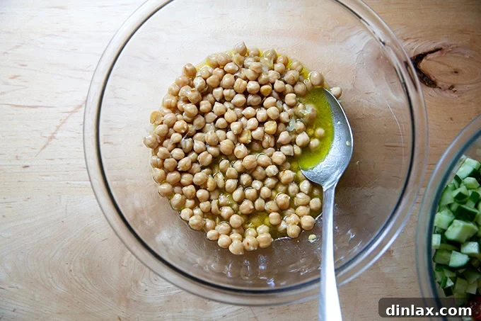A close-up of three cups of cooked chickpeas generously coated in a shimmering lemon vinaigrette within a large mixing bowl, ready for additional ingredients.