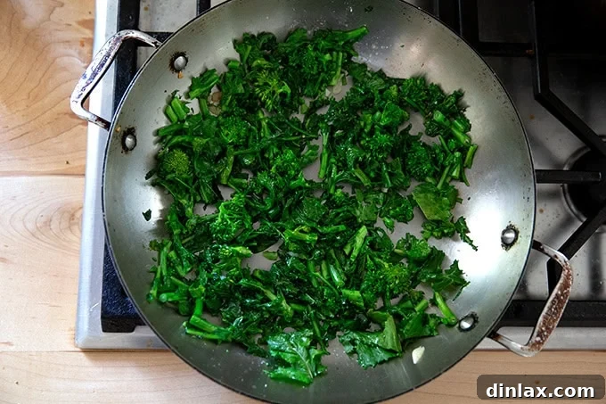 Freshly sautéed broccoli rabe, bright green and tender, in a large, hot skillet on a stovetop, ready to be added as a pizza topping.