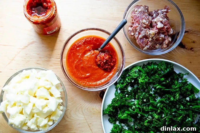 A selection of topping ingredients for a Grandma-style pizza: sautéed broccoli rabe, seasoned Italian sausage, shredded mozzarella, and a bowl of tomato sauce spiked with Calabrian chili paste, artfully arranged on a countertop.