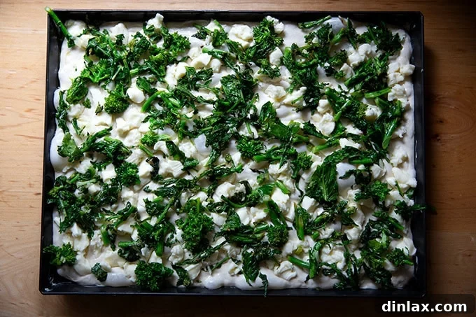 An unbaked Grandma-style pizza, beautifully topped with a generous layer of melted mozzarella cheese, followed by a vibrant spread of sautéed broccoli rabe, awaiting the sauce and final bake.