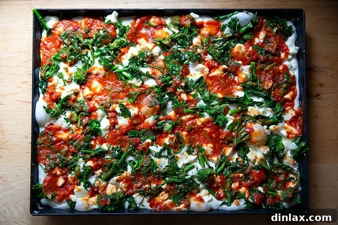 An unbaked Grandma-style pizza, artfully arranged with a base of cheese and sautéed broccoli rabe, now dotted with spoonfuls of vibrant tomato sauce, ready to be spread and finalized.