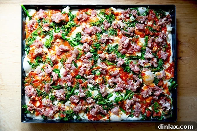 An unbaked Grandma-style pizza, fully assembled with layers of cheese, sauce, and sautéed broccoli rabe, now generously topped with small, savory pieces of Italian sausage, ready for the oven.