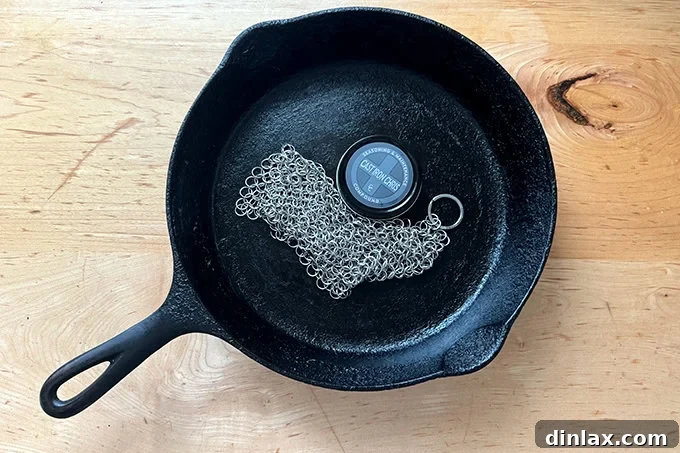 A well-seasoned cast iron skillet containing a Cast Iron Chris chainmail cleaner and a jar of seasoning wax, illustrating the essential tools for cast iron maintenance.